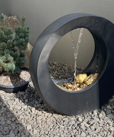 WASSERSPIEL IM LICHTHOF Kreisförmiger Brunnen mit Wasser, umgeben von Steinen und einer kleinen Pflanze.