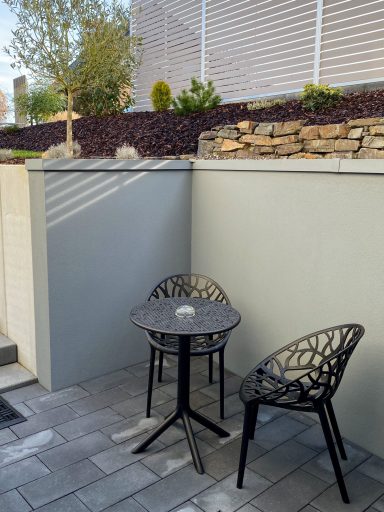 IM FREIEN Kleiner Tisch mit zwei Stühlen in einem Außenbereich, umgeben von Wänden und Pflanzen.