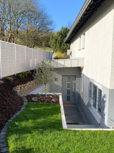 EINGANG FERIENWOHNUNG Haus mit modernem Eingangsbereich und gepflegtem Garten, umgeben von einem Zaun.